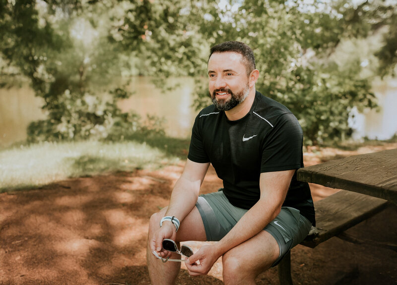 Natural dating app portrait of a man seated outdoors in Minneapolis, wearing athletic clothes and holding sunglasses