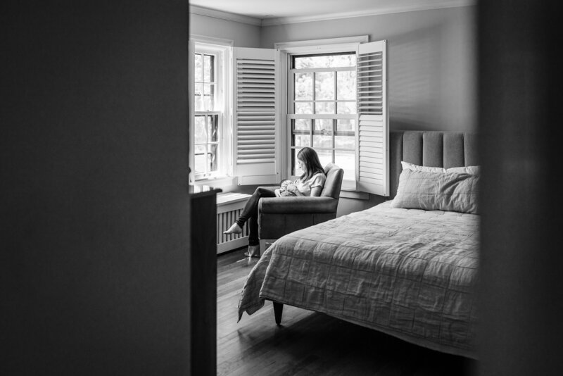 A wide-angle, black and white shot of a mother sitting in a cozy chair by the window, holding her newborn baby.