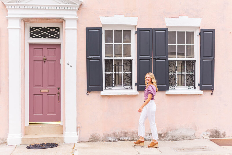 high school senior in purple boutique top and white jeans walking in front of purple historic home in downtown Charleston, South Carolina
