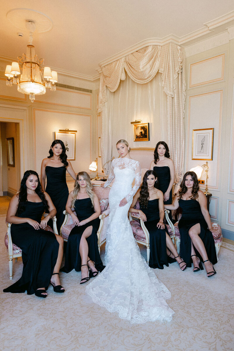 Bride in her gown posing with her bridesmaids that all wear chic black dresses.