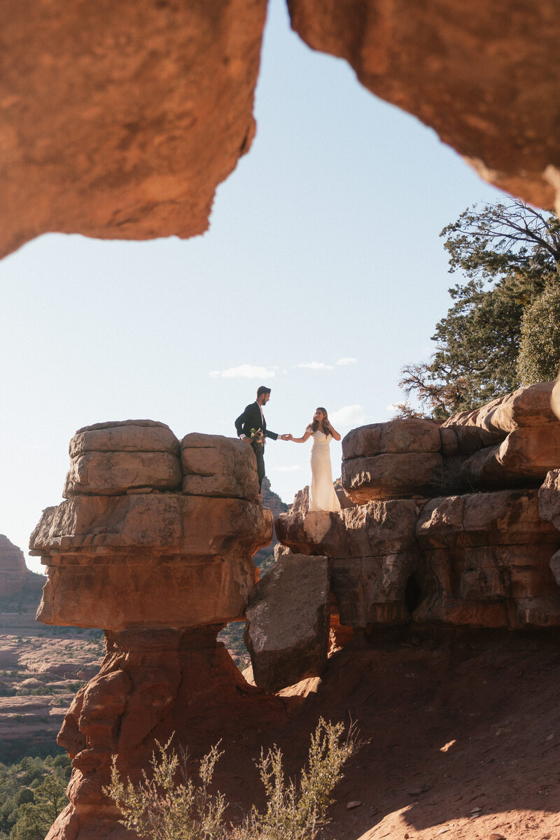 Bride and groom sharing a romantic moment on towering red rock formations at Merry Go Round Rock in Sedona during their adventurous elopement, surrounded by dramatic desert scenery.