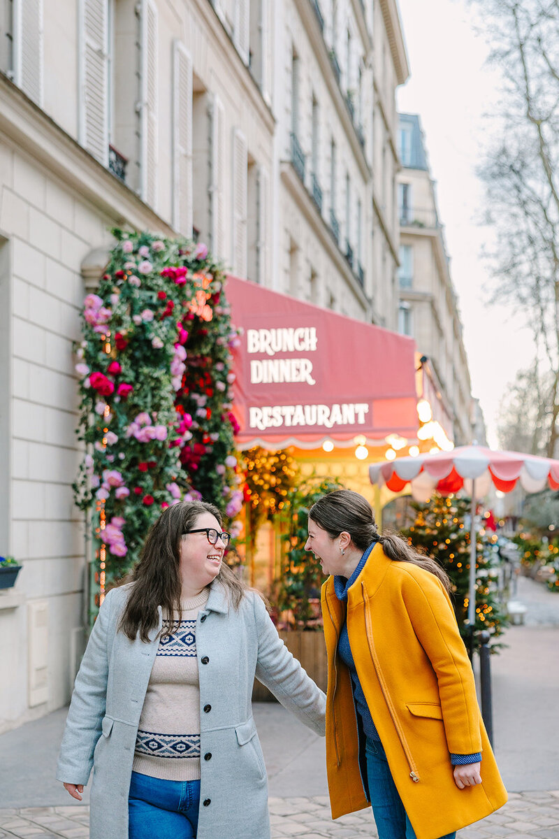 Two women laughing and holding hands while wearing jumpers and long colorful jackets during a Paris photoshoot near the Eiffel Tower on rue de l’Université.