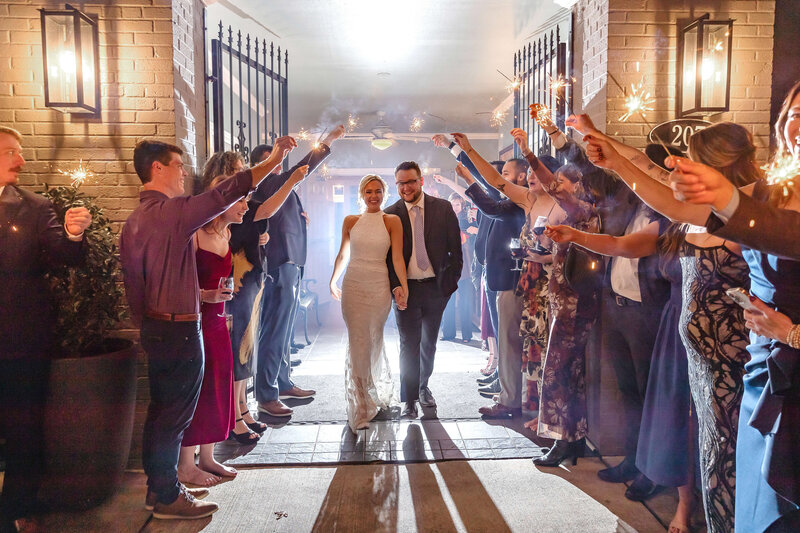 A bride and groom walk arm in arm through a tunnel of guests holding sparklers at night, smiling and celebrating their wedding. The scene is festive and joyful, surrounded by friends and family.