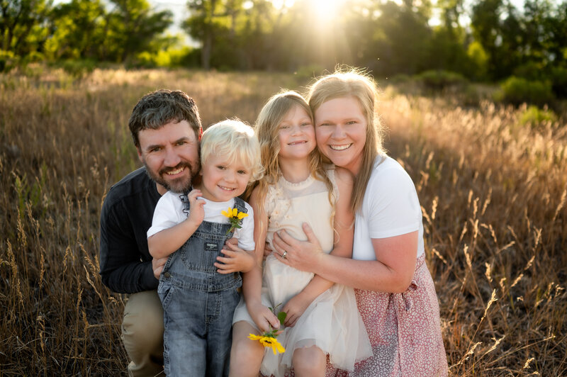 little boy in overalls holding a sunflower and smiling at the camera with his sister, mom and dad