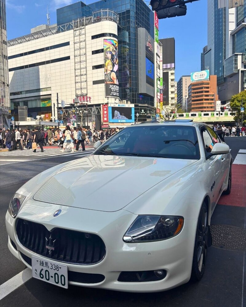 A sleek white Maserati Quattroporte paused dramatically in the middle of Shibuya Crossing, surrounded by bustling crowds and towering billboards.