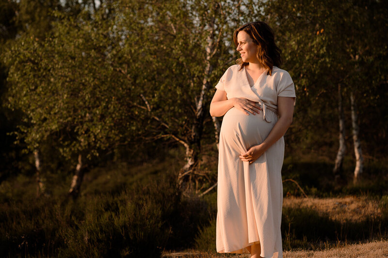 Zwangere vrouw staat in het licht van de zonsondergang met haar handen om haar bolle buik gevouwen en kijkt richting de zon