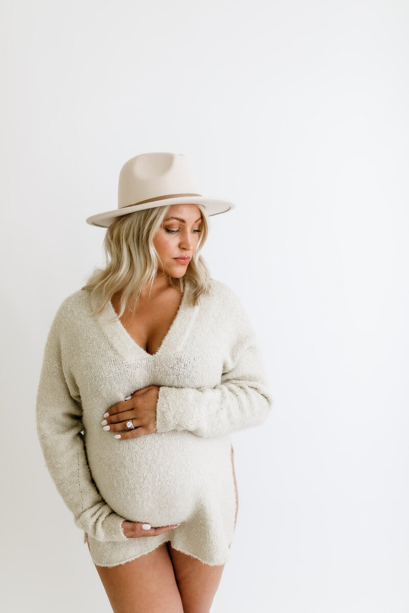 Pregnant woman in neutral sweater holding belly in all white studio