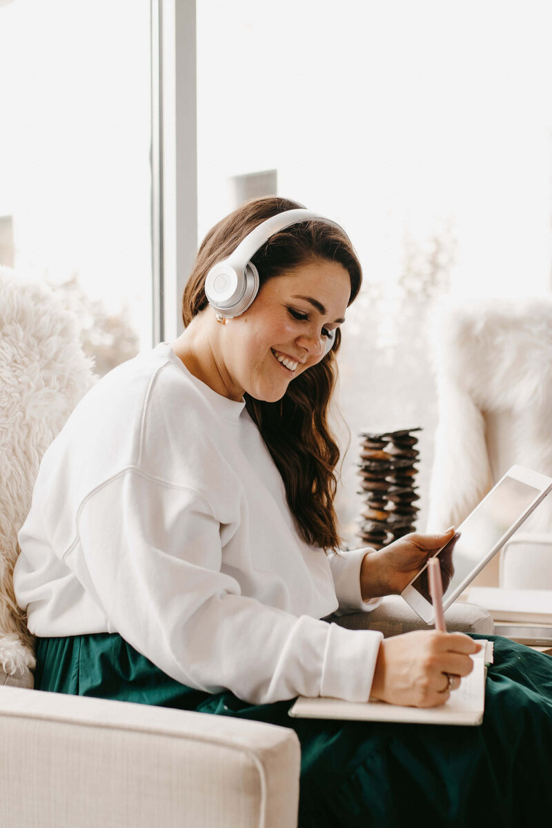 woman with dark hair white sweater teal skirt white headphones journaling and ipad