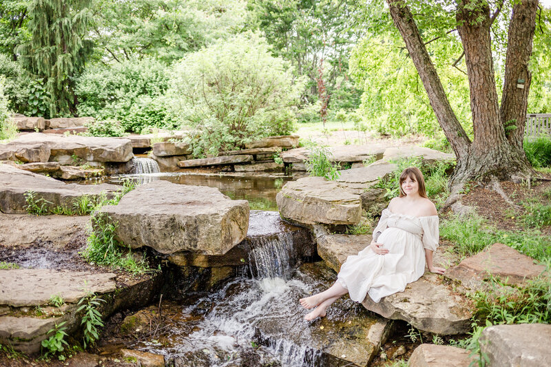 Mom to be wearing an off white shin length, off the shoulder dress sitting at a streaming waterfall at Cox Arboretum in Miamisburg, Ohio.