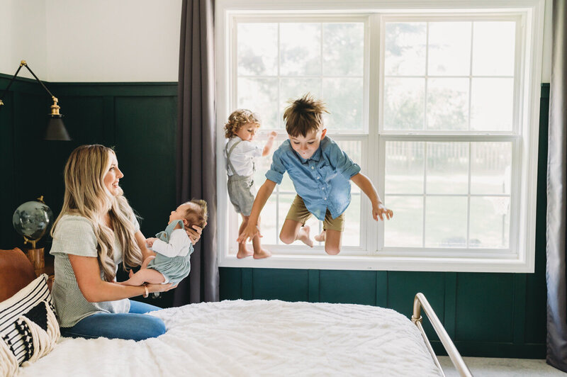 mom holding baby and older brothers jumping on bed