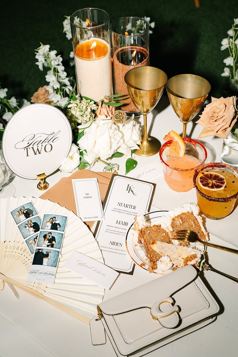 Wedding reception flat lay featuring a cake slice, photo booth pictures, menus, name cards, table numbers, gold champagne goblets, and signature drinks on the sweetheart table at Rancho Guijito Vineyard.
