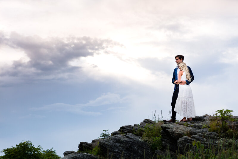 Newlyweds look out over the view from Cliffs of Glassy Chapel. He wears a casual blue suit and she wears a loose flowing sleeveless white dress with an intricate diamond pattern.