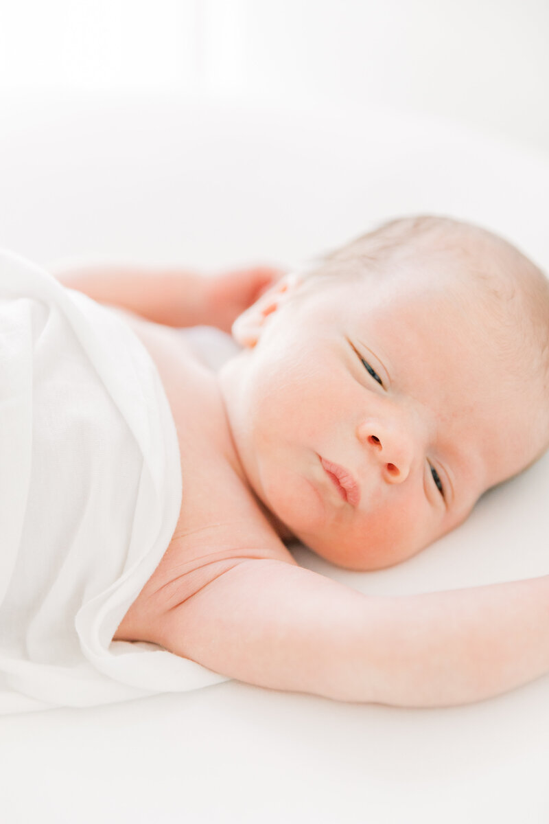 A newborn baby with sleepy eyes lays on a white bed under a blanket