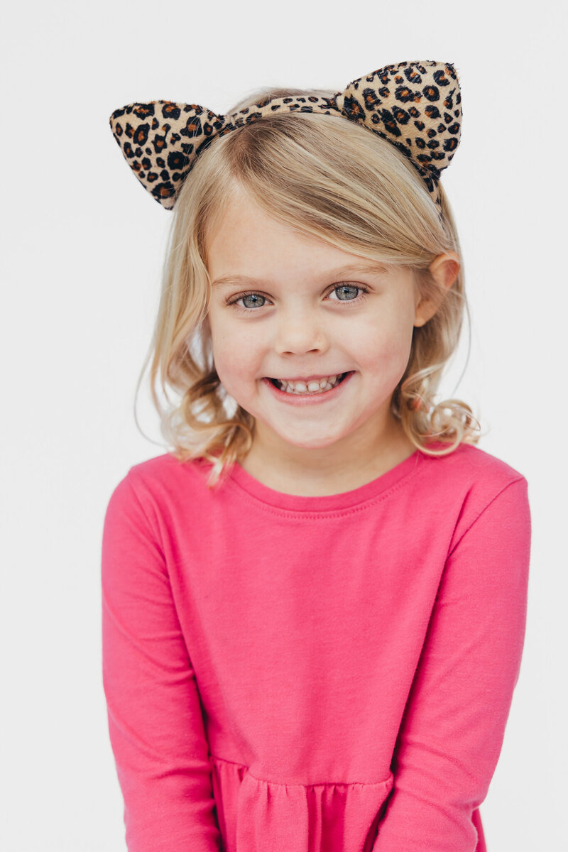 a blonde girl with leopard cat ears and a pink shirt smiles during her portrait session in San Diego with Christine Dammann