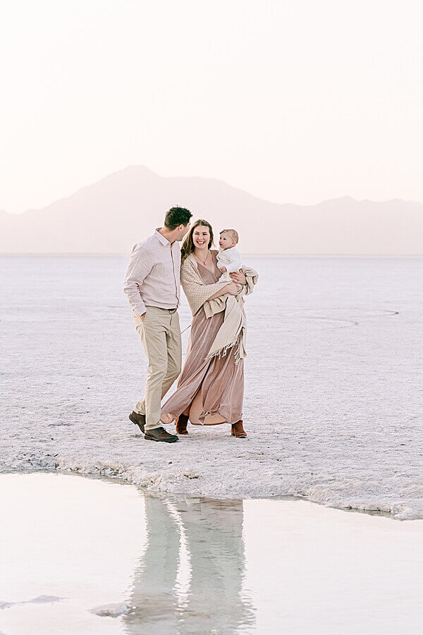 Family of three dressed in blush and khaki and white laughing at each other while they walk next to some water with mountains in the background. Portland family photography.