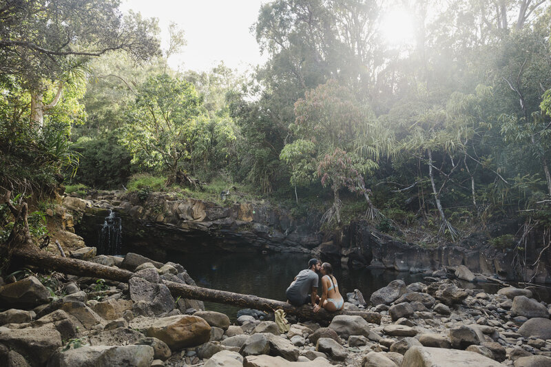 A couple sitting on the bank of a river kissing