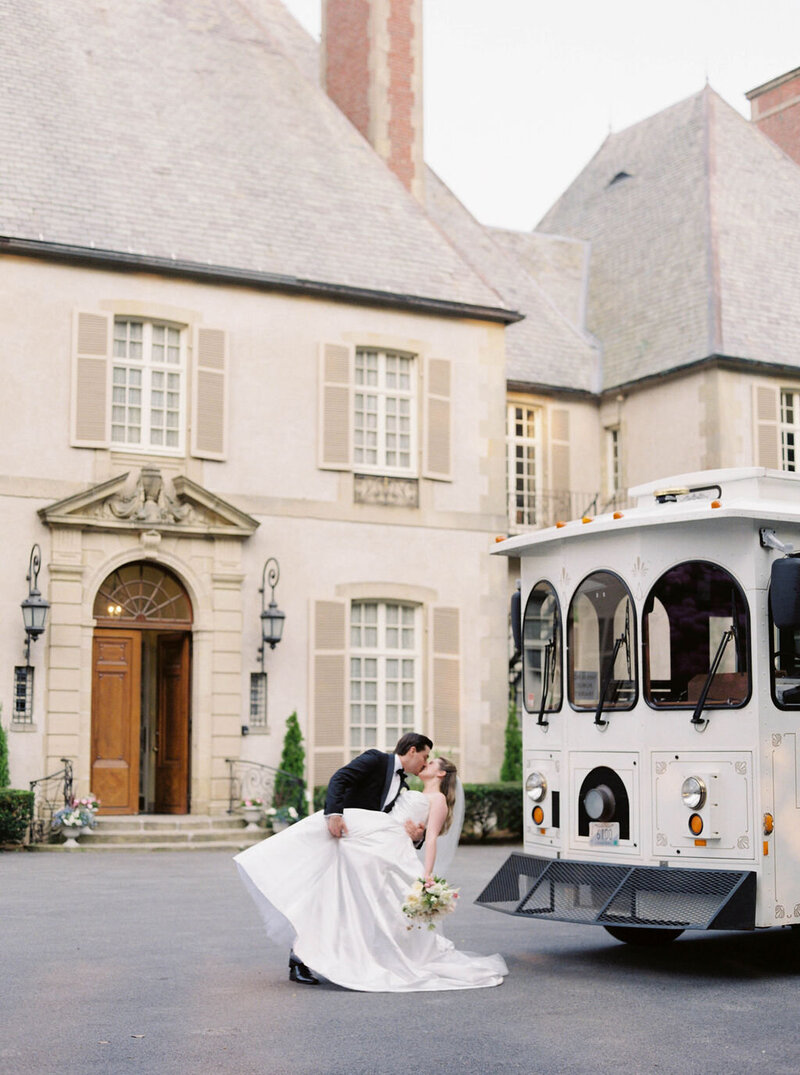 Rhode Island Wedding Photographer | A bride and groom share a romantic kiss in front of a grand, elegant mansion with a classic white trolley nearby.