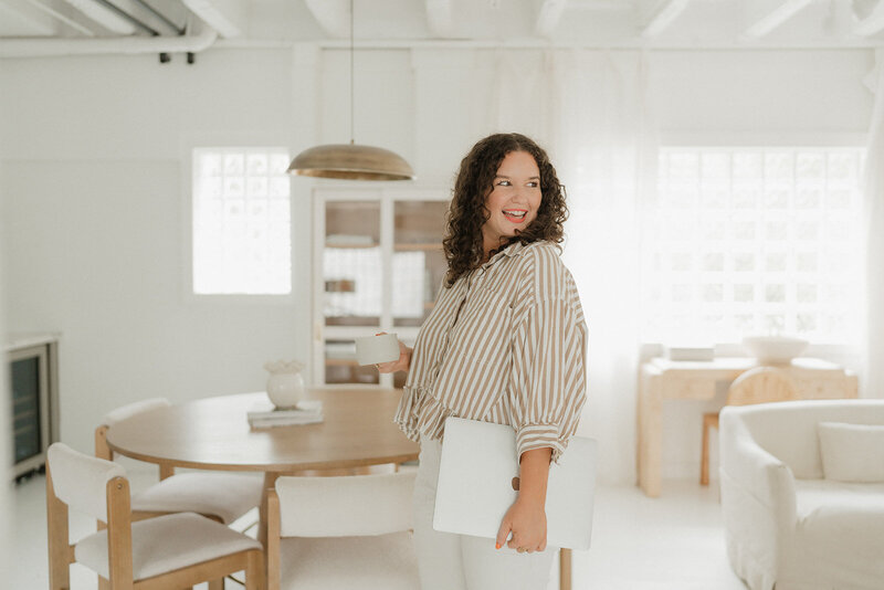 Morgan carrying a laptop and coffee cup by a table