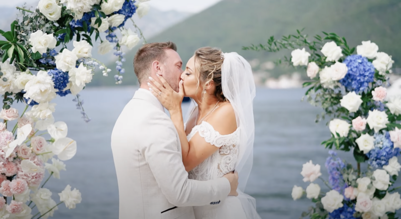 Bride and groom kiss in front of blue and blush flower arrangements with the Adriatic Sea behind them — romantic moment from a wedding video in Montenegro