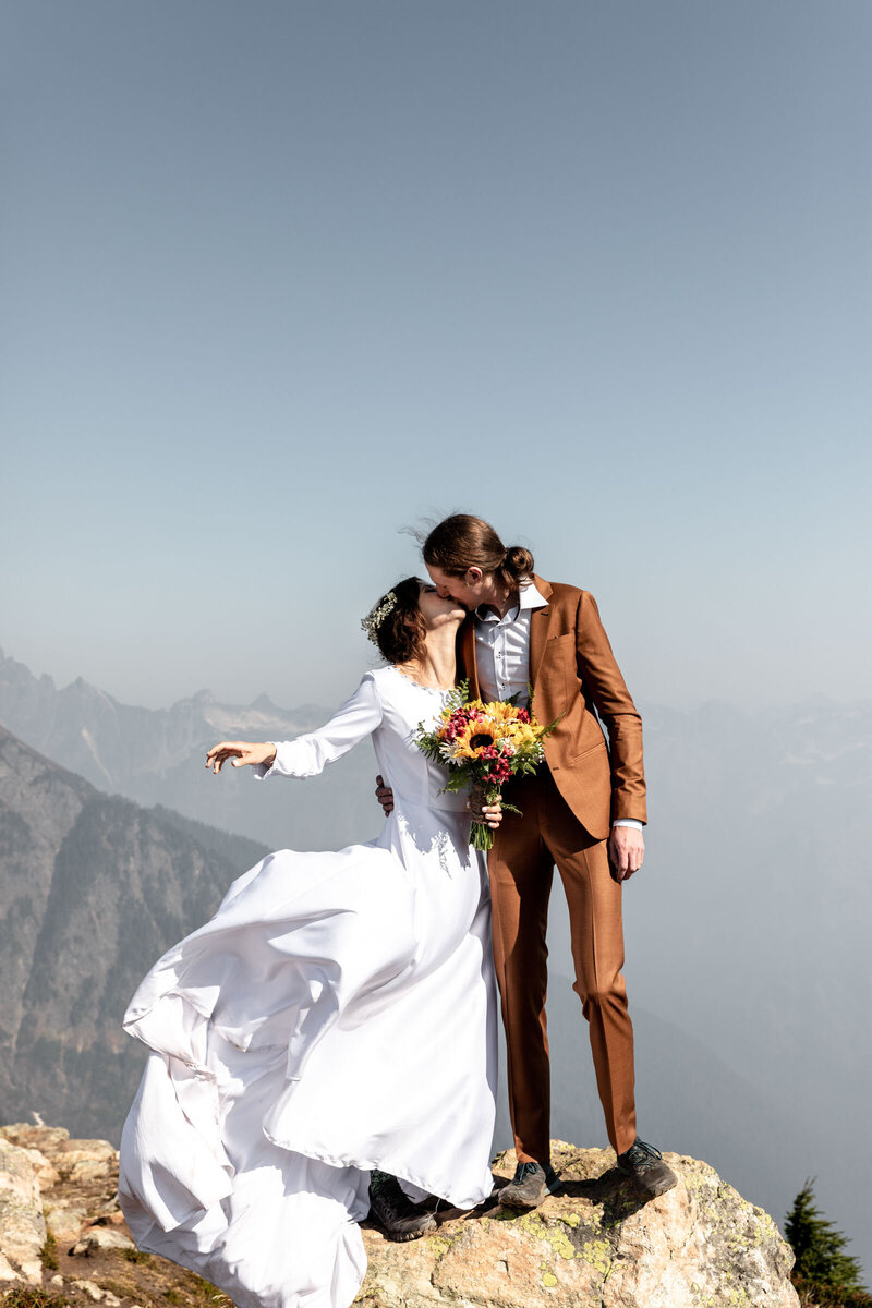 bride and groom are standing on a rock with a vast sky background and mountains behind them. They are kissing and her white dress is blowing in the wind. She is holding a bouqet with yellow sunflowers