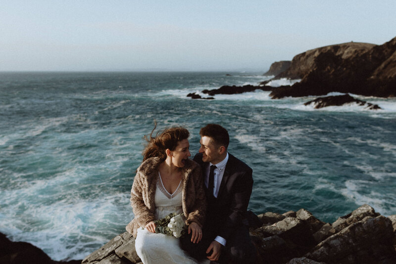 bride and groom sitting on rock near water