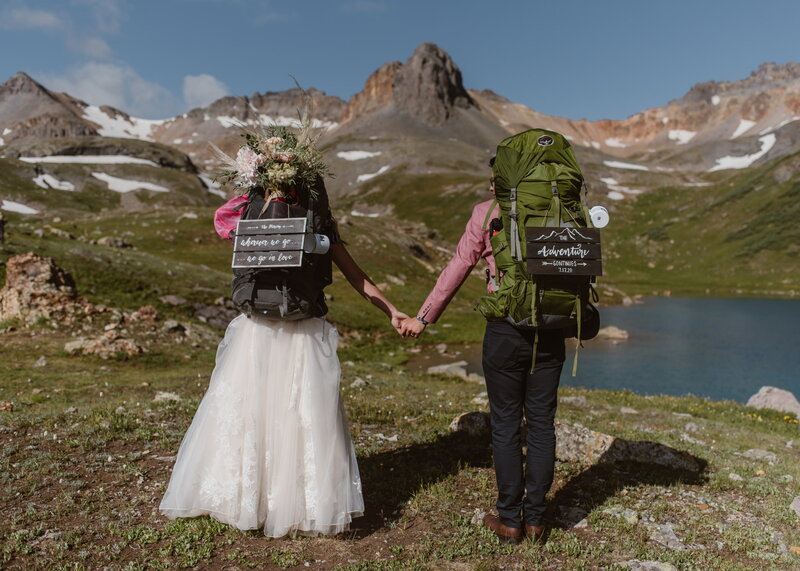 Colorado backpacking elopement photographer captures a bride and groom holding hands with hiking backpacks beside an alpine lake in the San Juan Mountains.