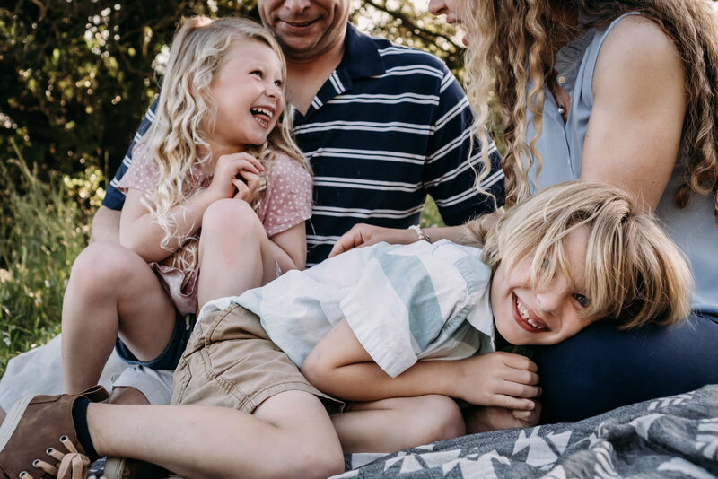 a family of 4 sits on a blanket in the grass with trees behind them.  A blonde haired boy in light clothing with red lips and blue eyes lays with his head in his mothers lap and stares at the camera with a big smile.  His younger sister is sitting in his fathers lap behind him laughing with her tongue out.  She las long white blonde curlse and as soft pink polk-a-dot shirt.