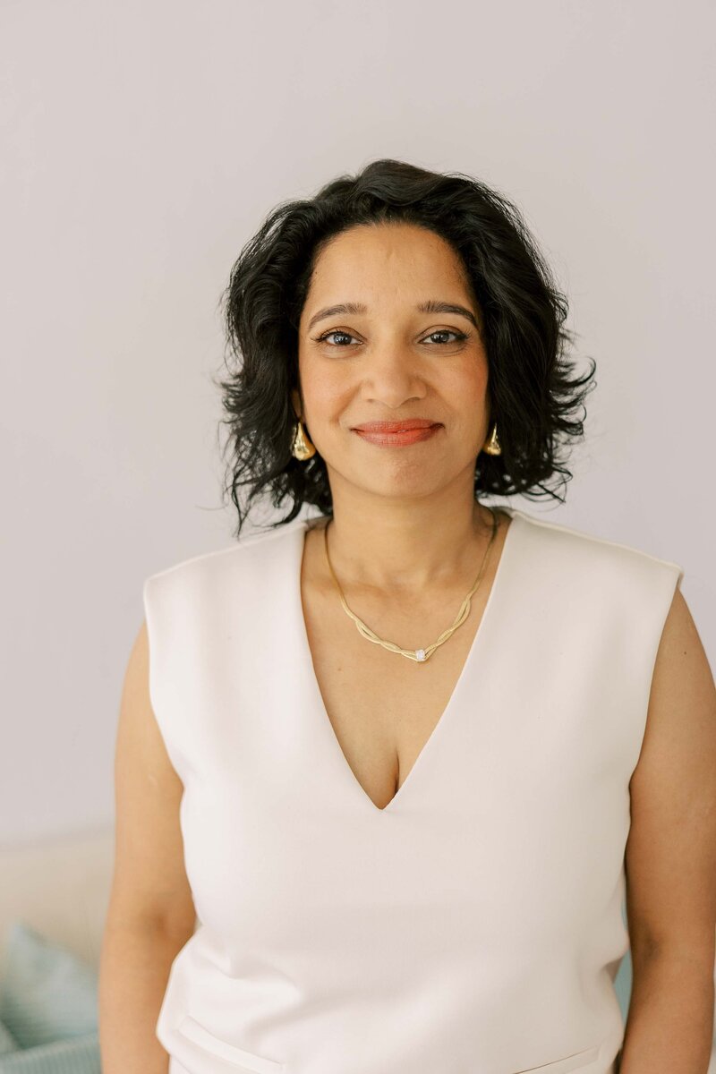 Woman wearing gold jewelry and an all white outfit and smiling at the camera with a soft smile