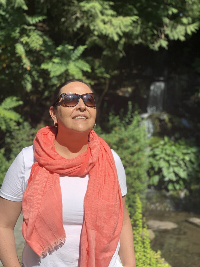 Marita Choque standing peacefully in nature near a waterfall, wearing a coral scarf and sunglasses, symbolizing renewal and grounded energy.