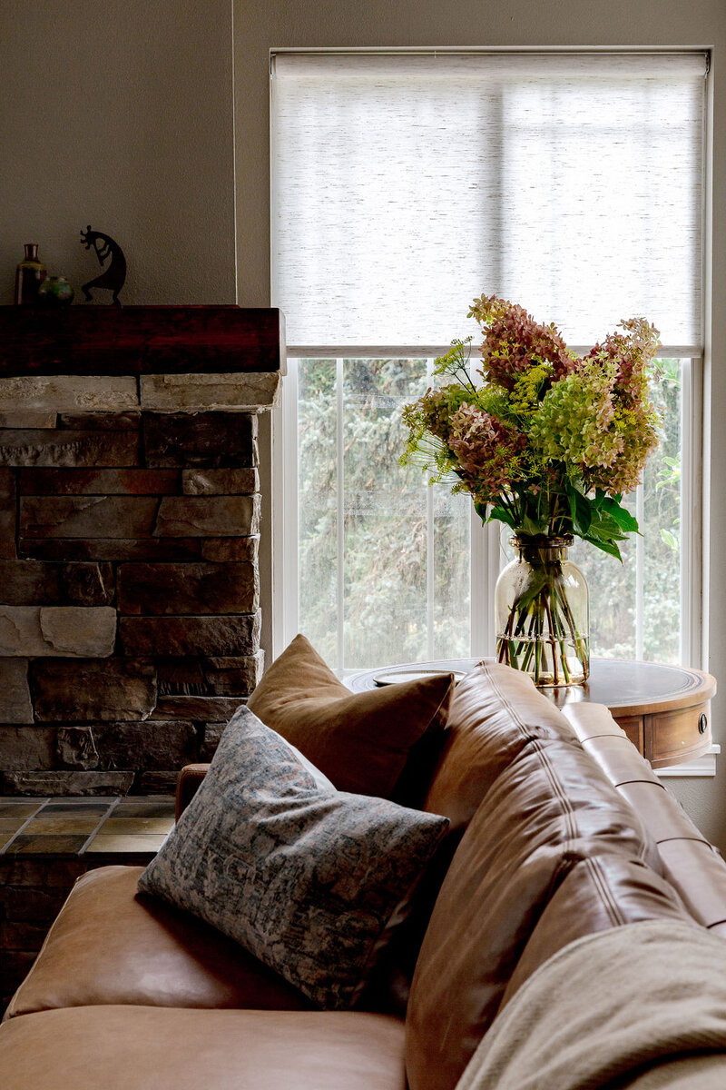 A living room with a brick fireplace, leather couch, side table in front of windows.