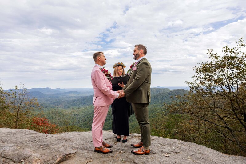 Two grooms stand facing each other holding hands during their elopement ceremony. There is an officiant in the background dressed in black and wearing a flower crown. They are standing on a large rock overlooking the vast mountains.