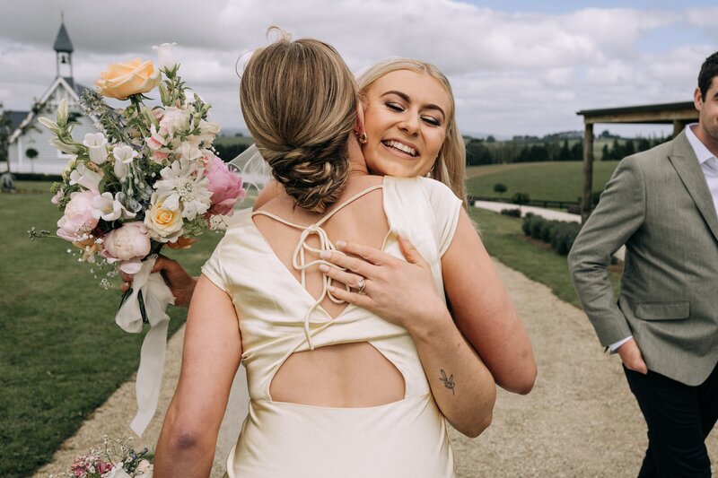 bride hugs bridesmaid in pale yellow dress at the farm chapel wedding venue in waimumu