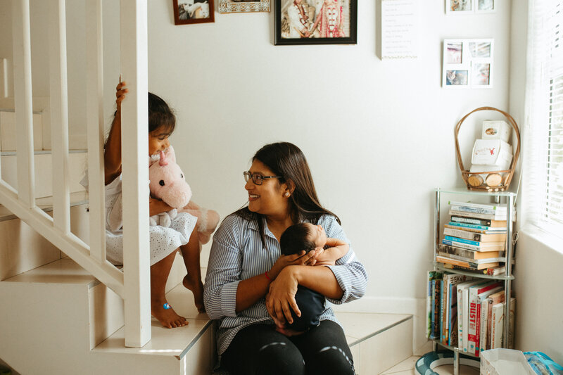 A mother holds her baby at the bottom of a staircase while her toddler clutches toys and and giggles at her new baby sibling. Baby Photos by Rebecca Rottcher