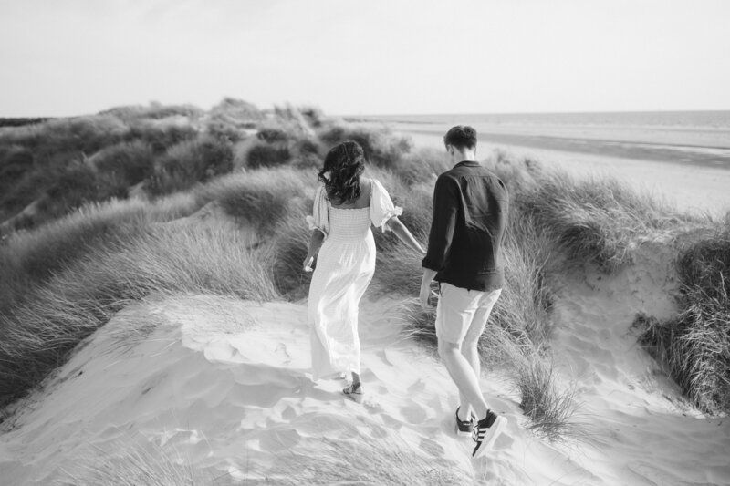 A black-and-white pre-wedding shoot captures a couple walking hand in hand over the sand dunes at West Wittering. The woman wears a long dress, and the man is in shorts and a shirt. Tall grass sways gently as the beach stretches out in the background.