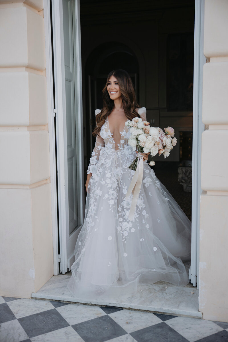 Bride in a stunning Berta wedding dress holding a bouquet of white and blush flowers, standing at the entrance of Villa Astor.