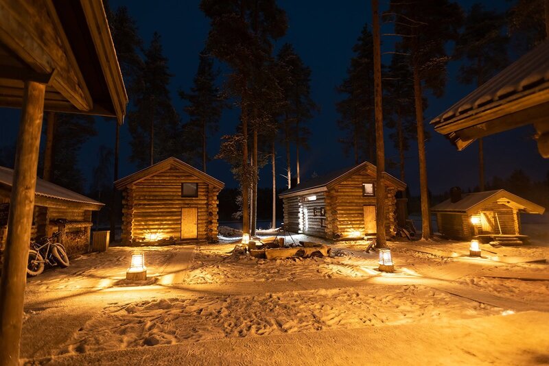 Three quaint wood cabins during an adventurous luxury elopement in the Arctic Circle stand bathed in warm light during the night.  The ground is covered in snow and in the dark blue night sky, the shadow of tall straight pine trees is lurking. 