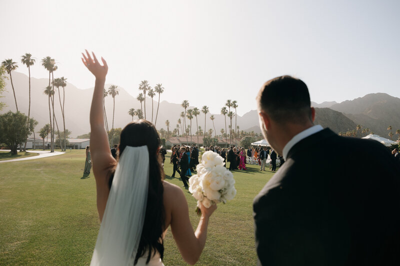 Photo from behind of bride and groom walking towards their reception with their guests in the background