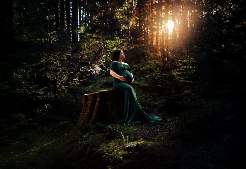 Pregnant woman wearing an off shoulder ruffle green dress sitting on a tree stump in the forest at Buntzen Lake Anmore with sunset light shining through the trees.