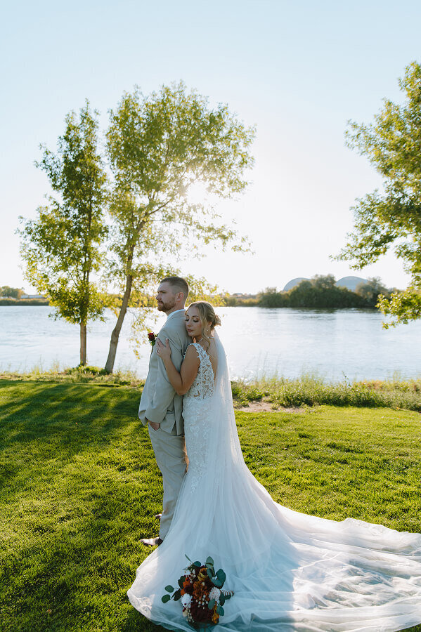 A bride lovingly holds onto her groom's chest from behind. He's looking candidly to the side. They're standing in grass with a large lake behind them in Idaho Falls, ID.