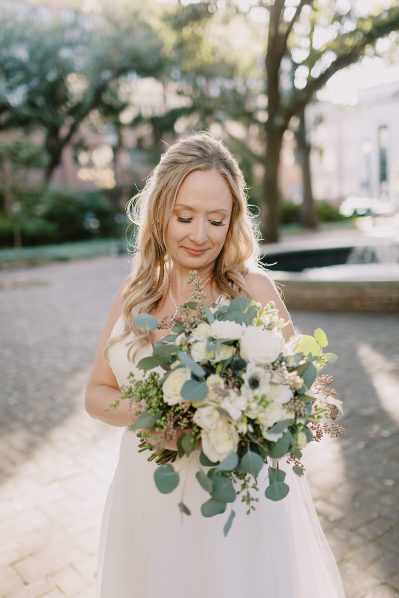 Bride looks down at floral bouquet of eucaluptus and white florals backlit by the sun and stands on a cobble stone sidewalk in front of a water fountain.