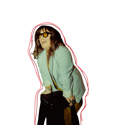 Women wearing sunglasses and a blue suit jacket, laughing and smiling while leaning on a stool