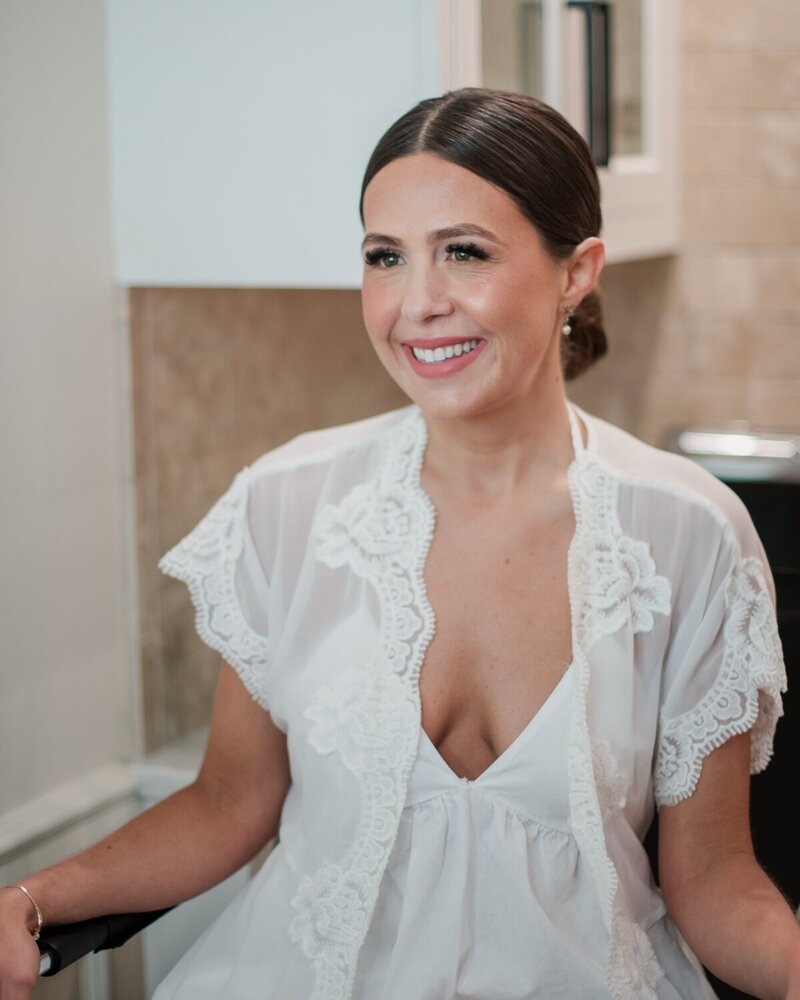 Bride in a white lace robe smiling during wedding prep, featuring natural glam makeup with soft pink tones—created by Artistry by Jessica.