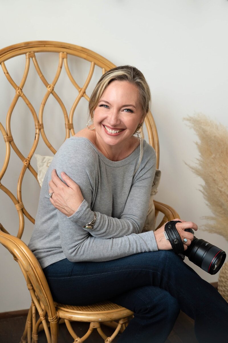 Headshot of Jen Denton sitting in a grey shirt and smiling holding a camera