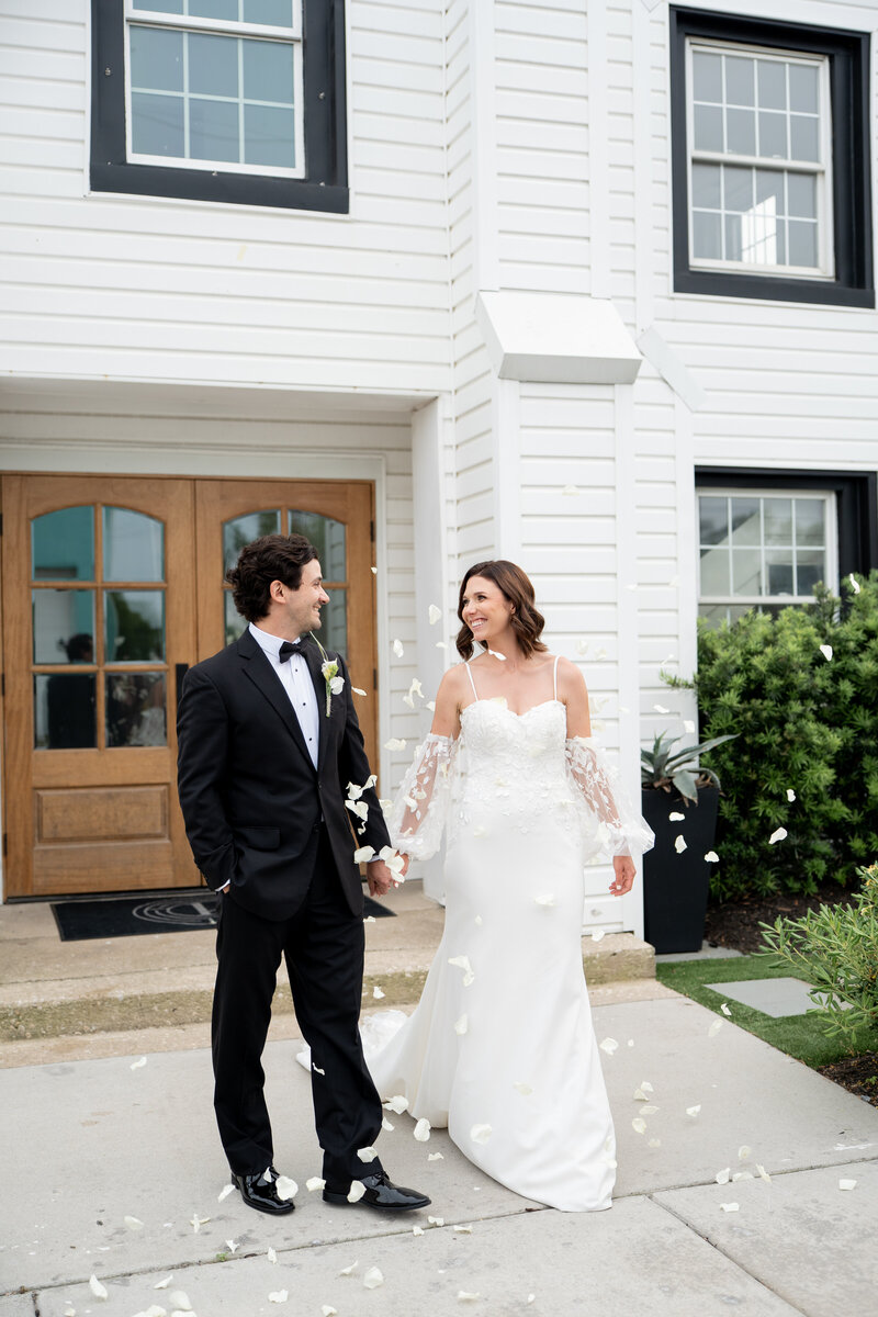 A newly married couple exiting the ceremony. They are happy and smiling at each other. White flower petals fall around them 
