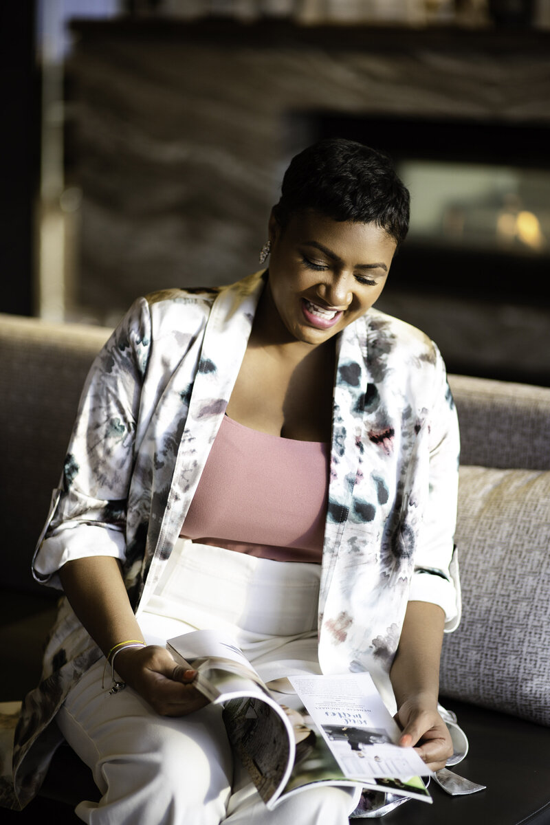 Alicia Mae reads a magazine on a sofa. She is wearing a pink shirt, a white/floral cardigan, and white pants