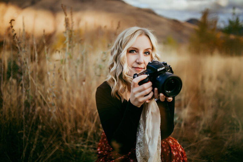 Woman with blonde curly hair crouched down and holding a camera and smiling.