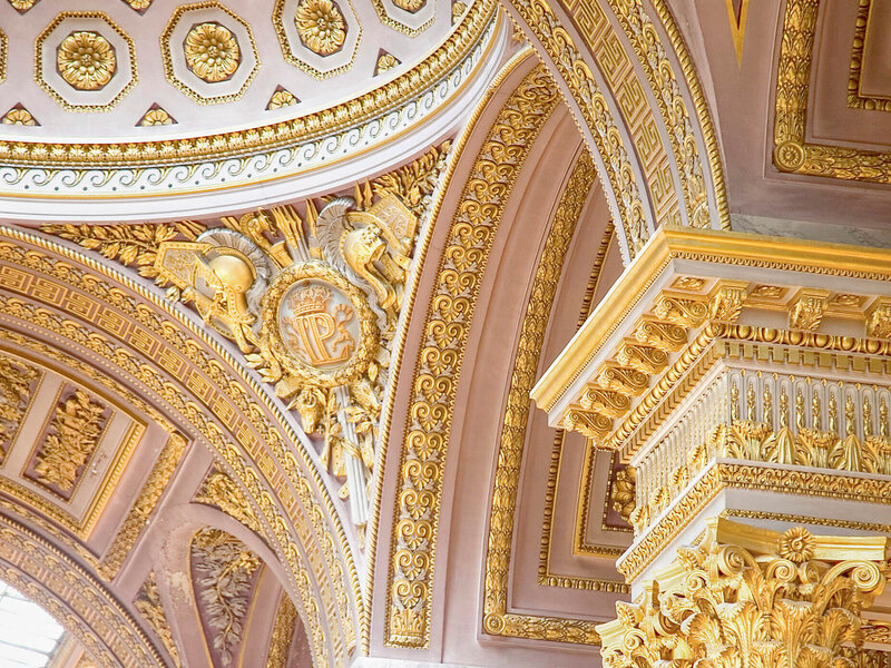 Ornate architectural details featuring gold and cream decorative elements on a ceiling and arch. The design includes intricate patterns, medallions, and lavish embellishments with a regal and luxurious appearance.