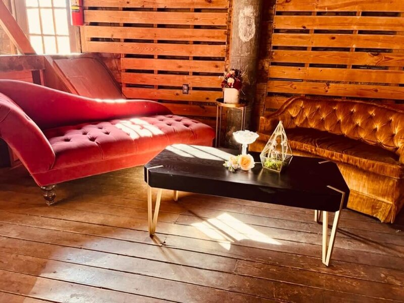 A rustic room with wooden walls and floors features a red chaise lounge, a brown couch, and a black coffee table with gold legs. The table holds a geometric terrarium and decorative items. Sunlight streams through a nearby window.
