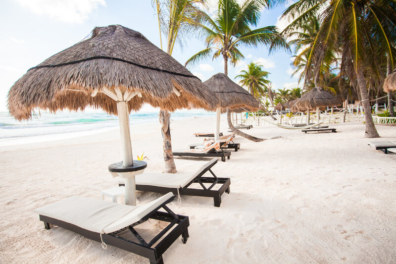 Escape to a tropical paradise featuring palm trees, straw umbrellas, and wooden lounge chairs on pristine white sand. Perfect for Caribbean getaways, the ocean waves gently roll under a partly cloudy sky, offering an idyllic setting for your luxury vacation dreams.