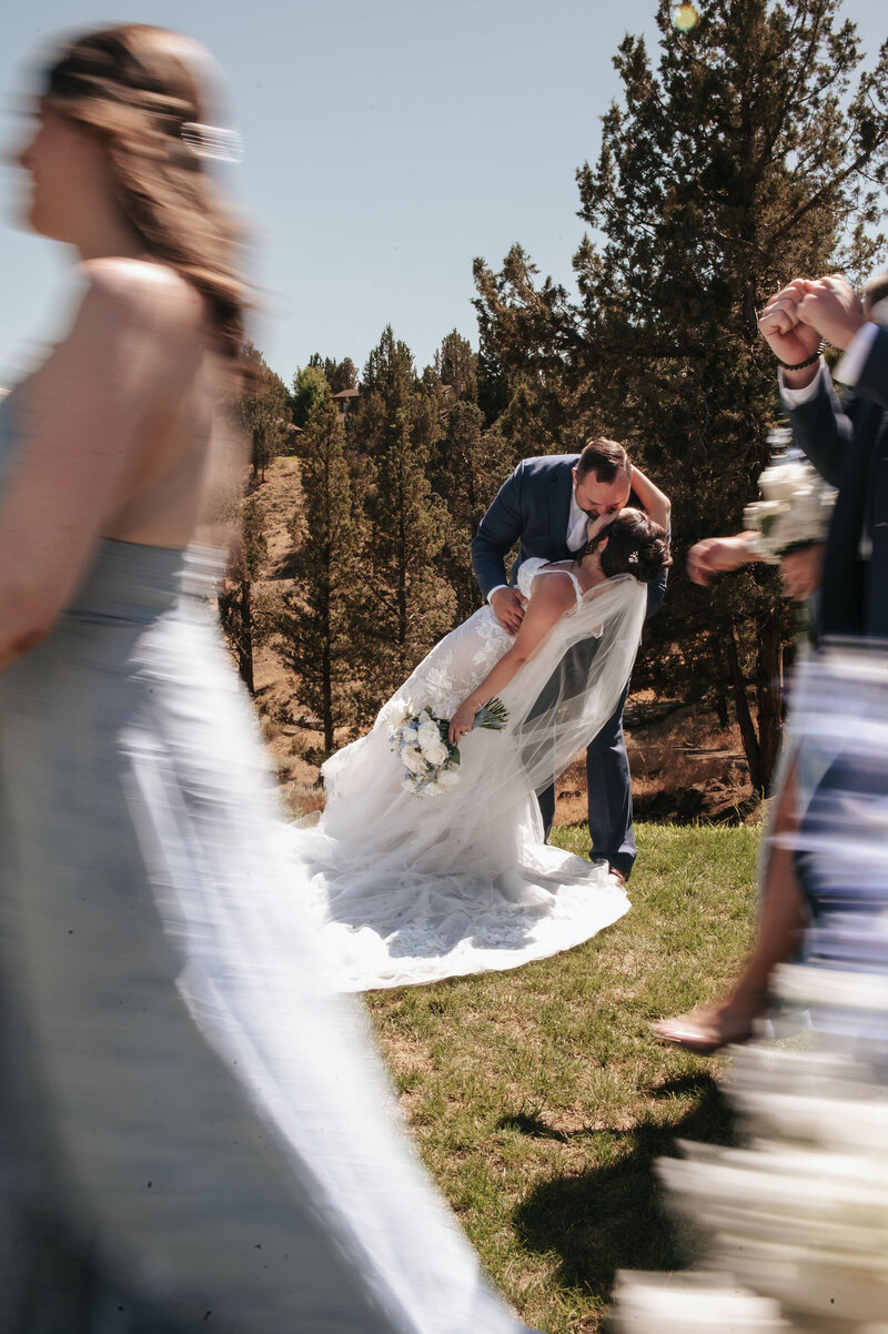 Romantic wedding moment at River Run Lodge as a couple shares a dip and the bride’s dress sways gracefully in motion, with the bridal party walking by in a soft blur in an outdoor setting.